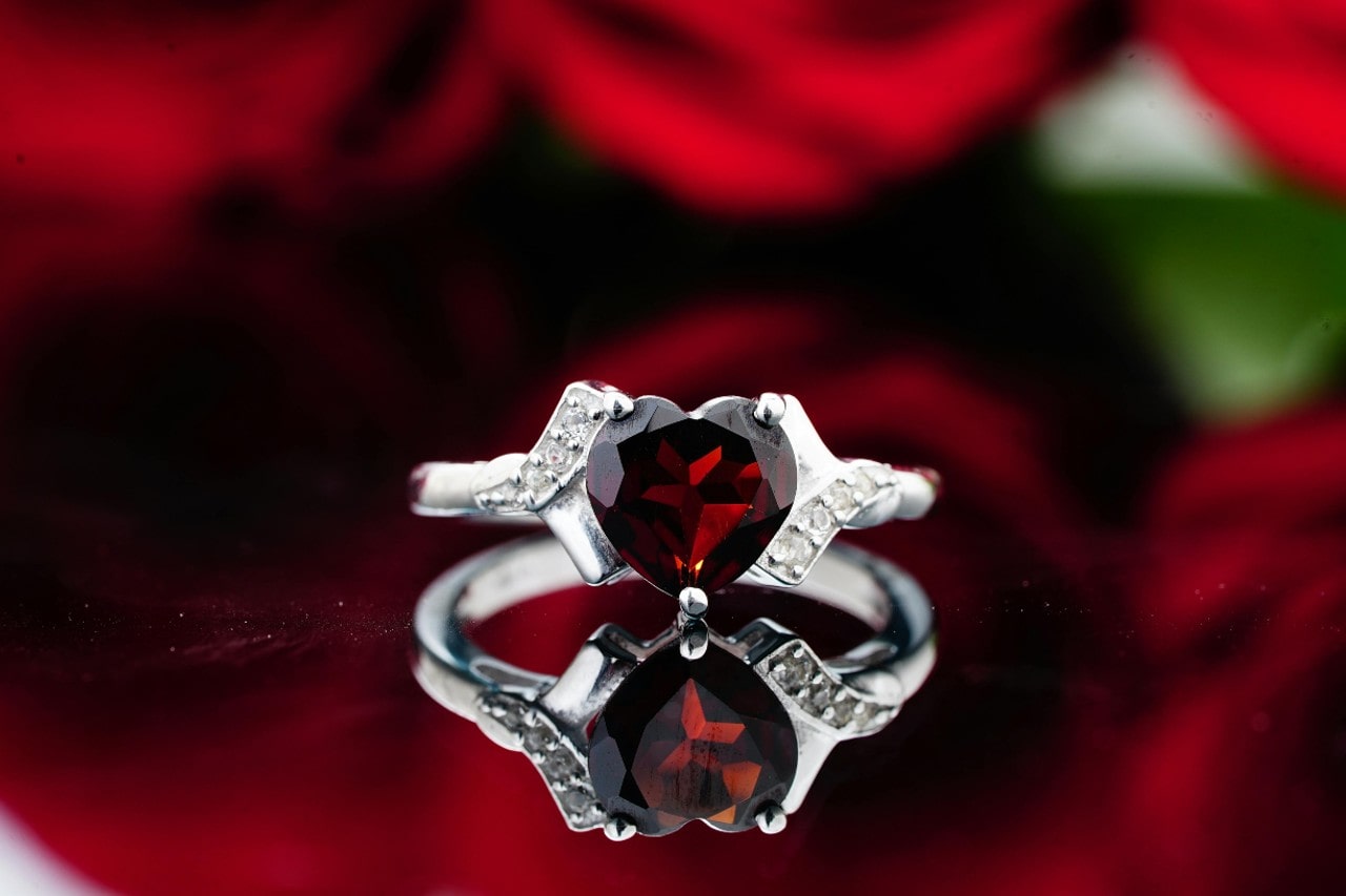 A close-up of a stunning ruby engagement ring displayed before a bouquet of red roses on a reflective surface.