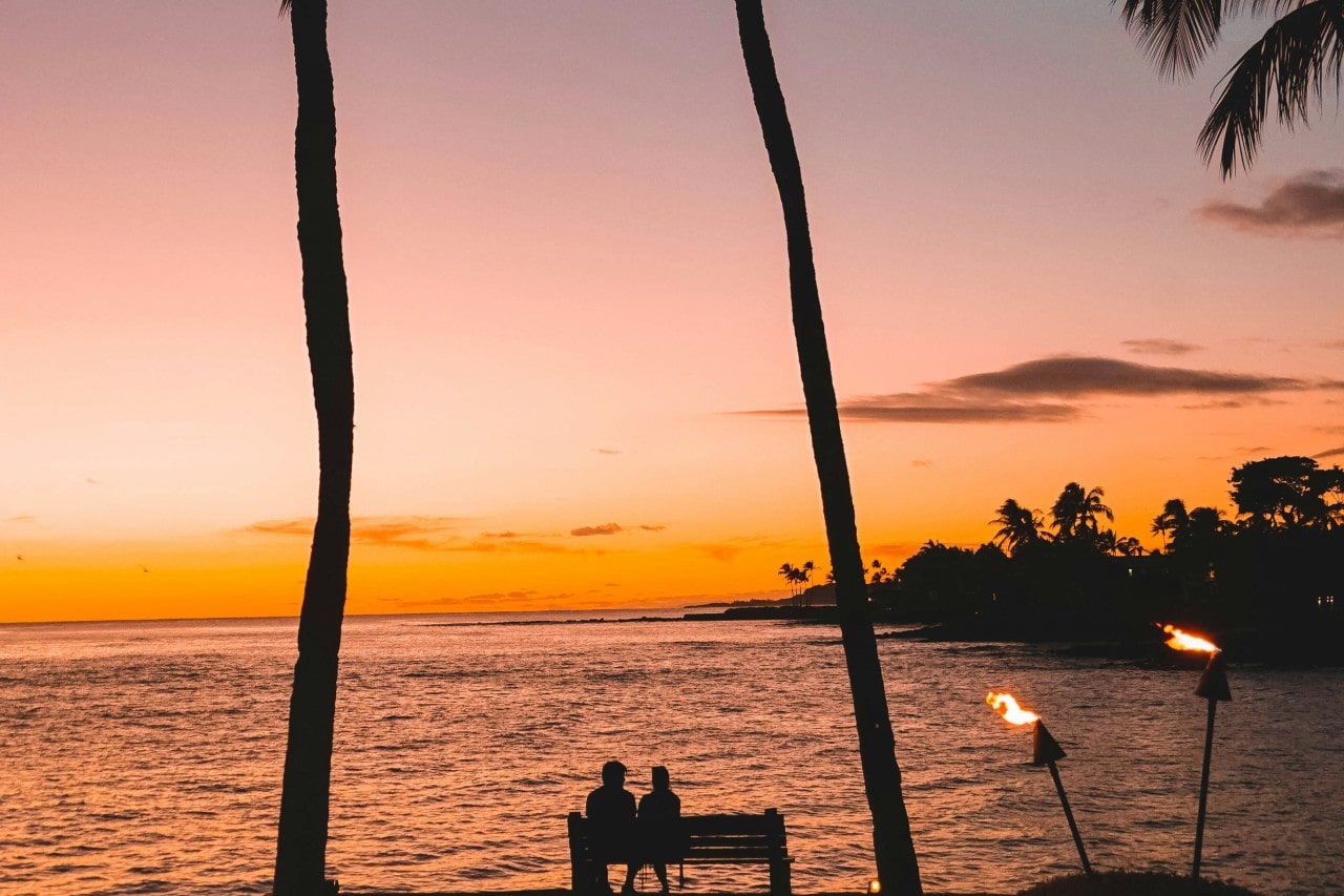 An engaged couple sitting at a waterside bench as the sun sets.