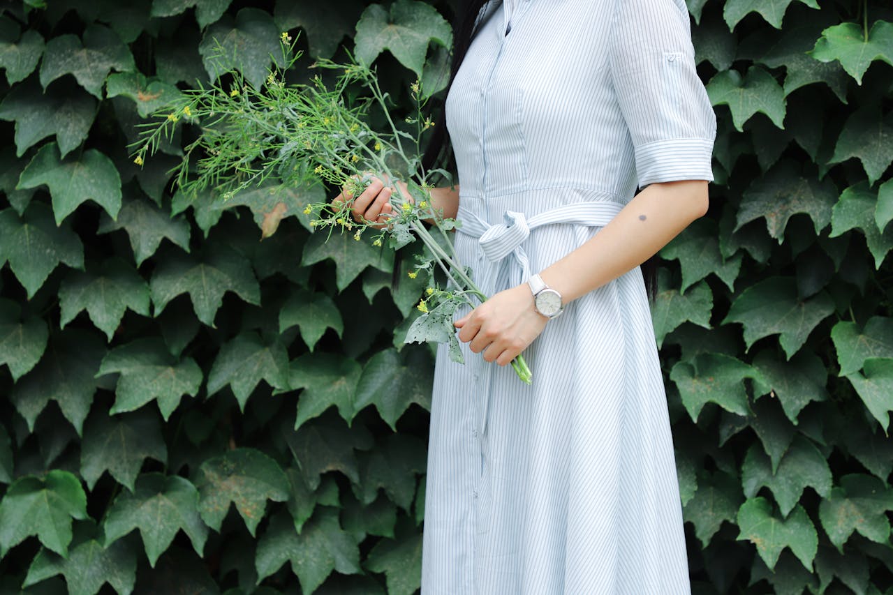 A shot of a woman wearing a luxury watch, standing in front of a wall of ivy and holding a few plant sprigs.