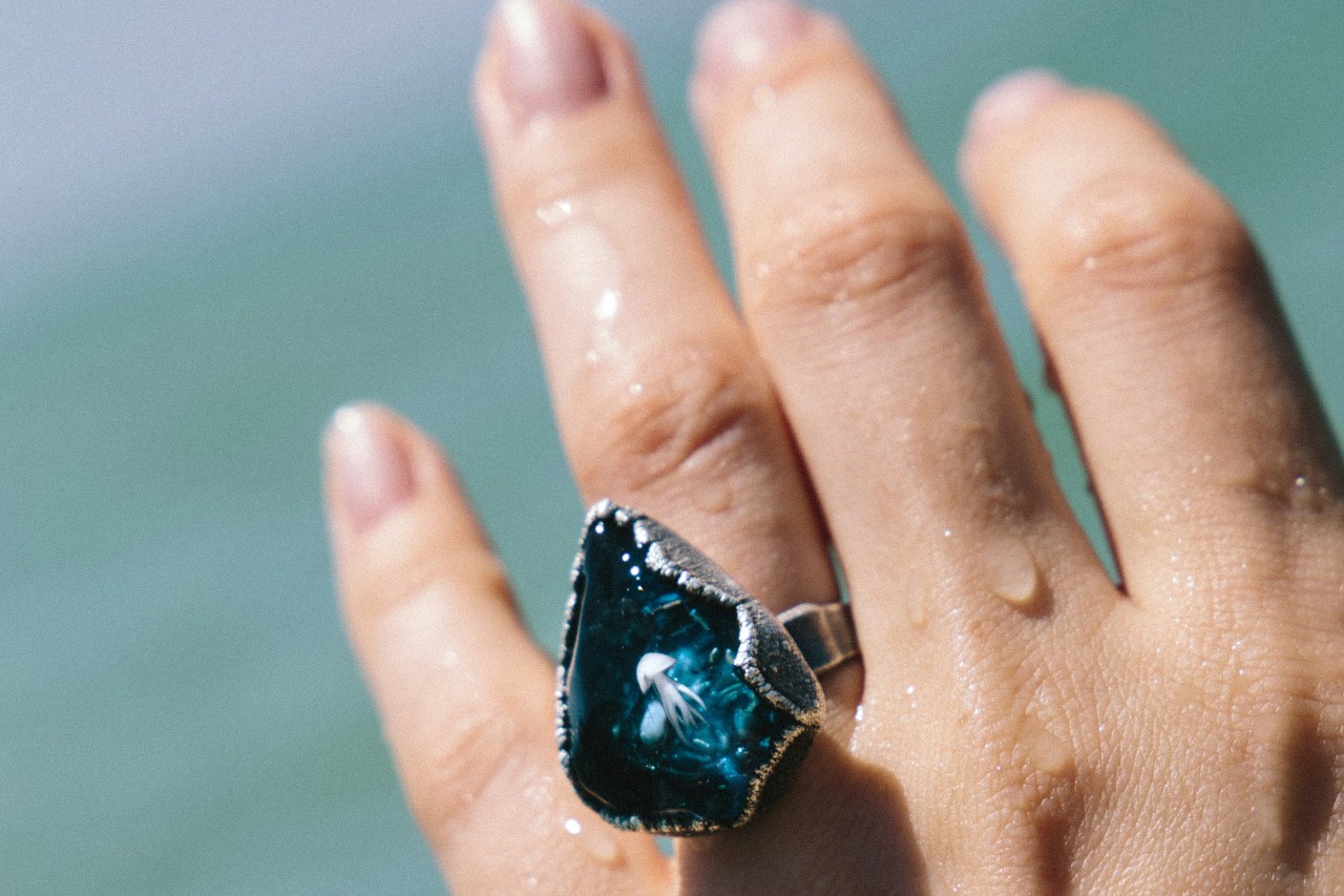 A woman’s hand in front of the ocean, adorned with a white gold fashion ring with a large blue stone.