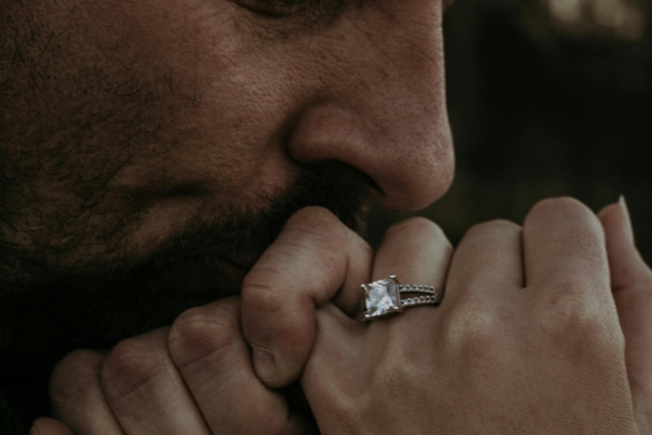 a man kissing a woman’s hand, which is adorned with a princess cut engagement ring