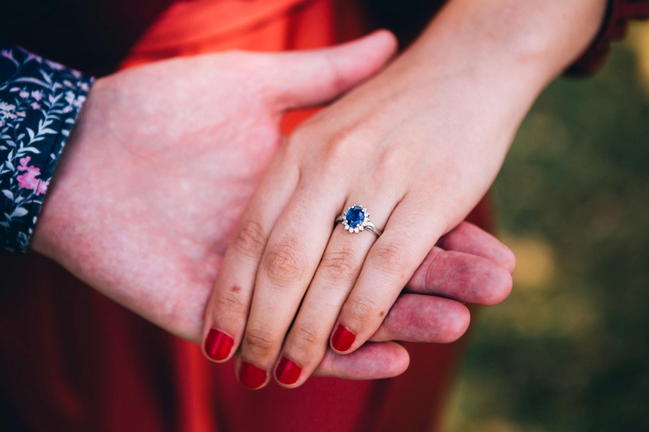 a man’s hand holding a woman’s, whose hand is adorned with a sapphire engagement ring