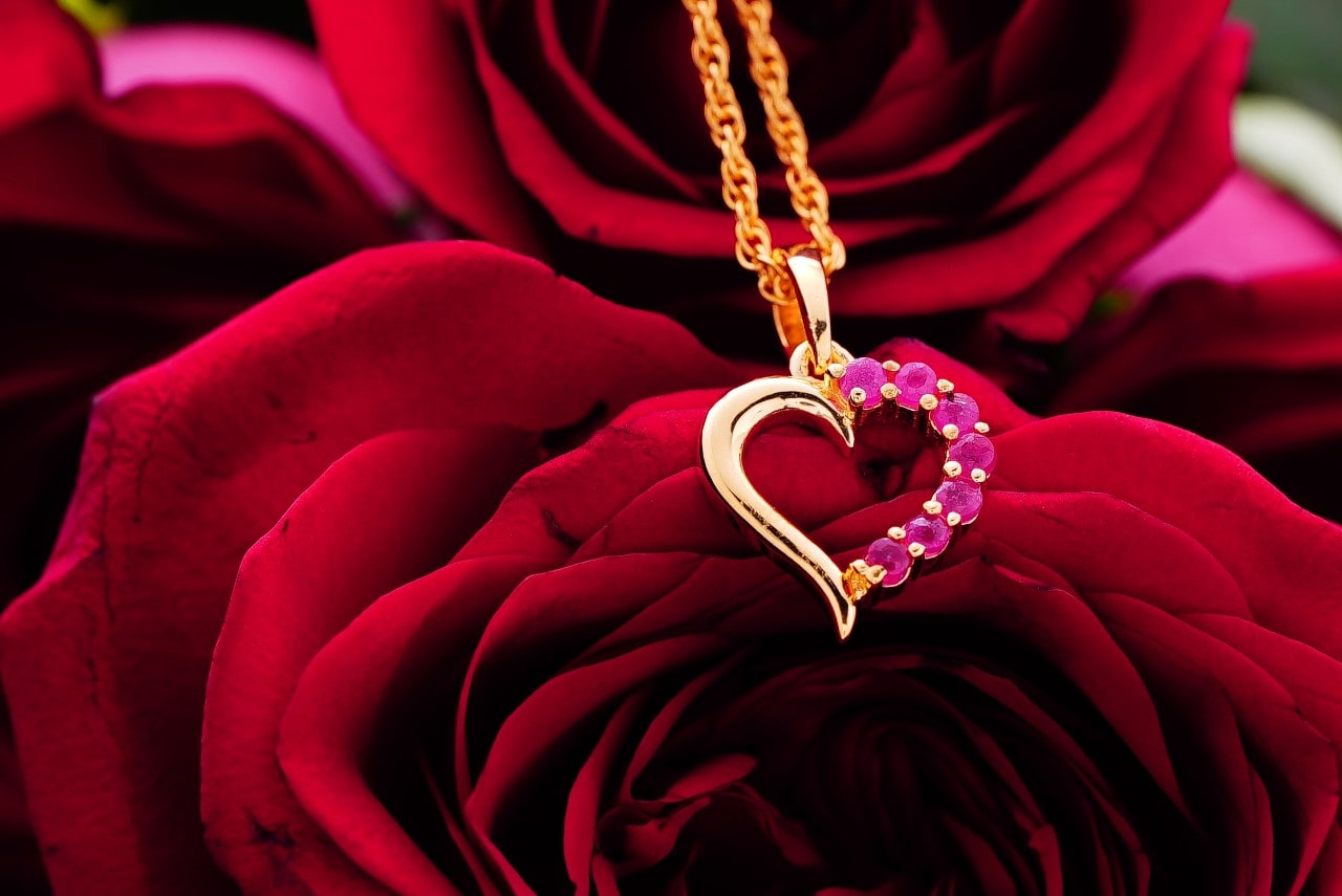 A yellow gold and ruby heart necklace resting on a red rose