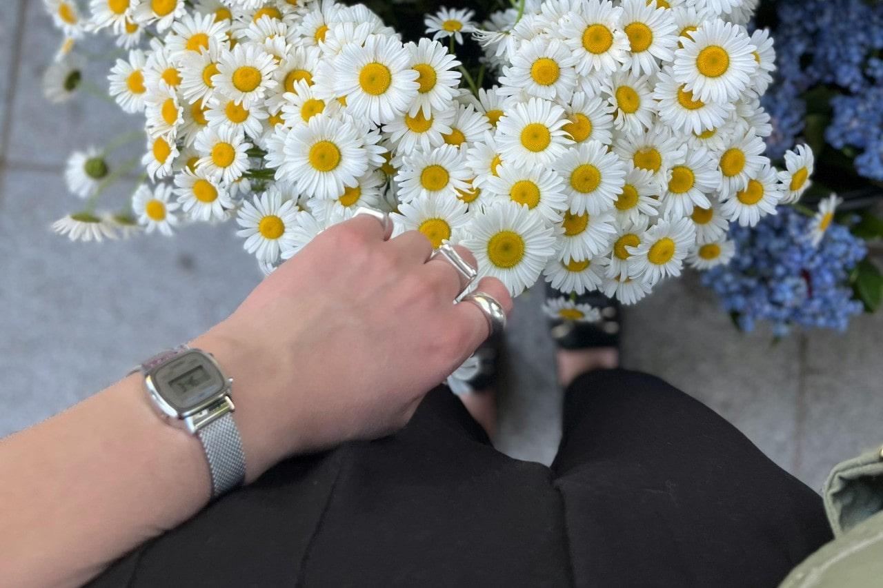 a person wearing a silver watch standing near a bunch of daisies