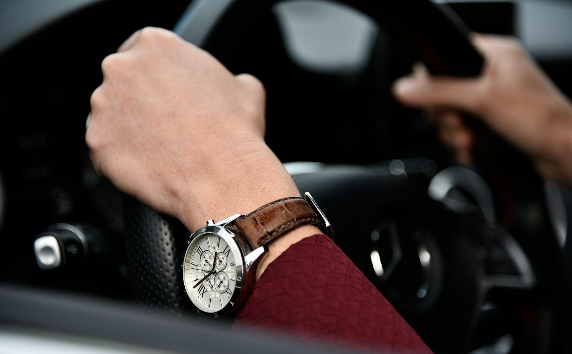 a person’s hands holding a steering wheel and wearing a watch with round case a leather strap.