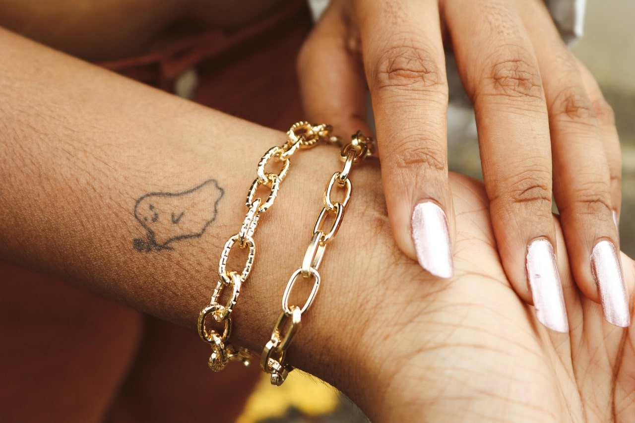 A woman with a ghost tattoo wearing two chunky gold chain bracelets.