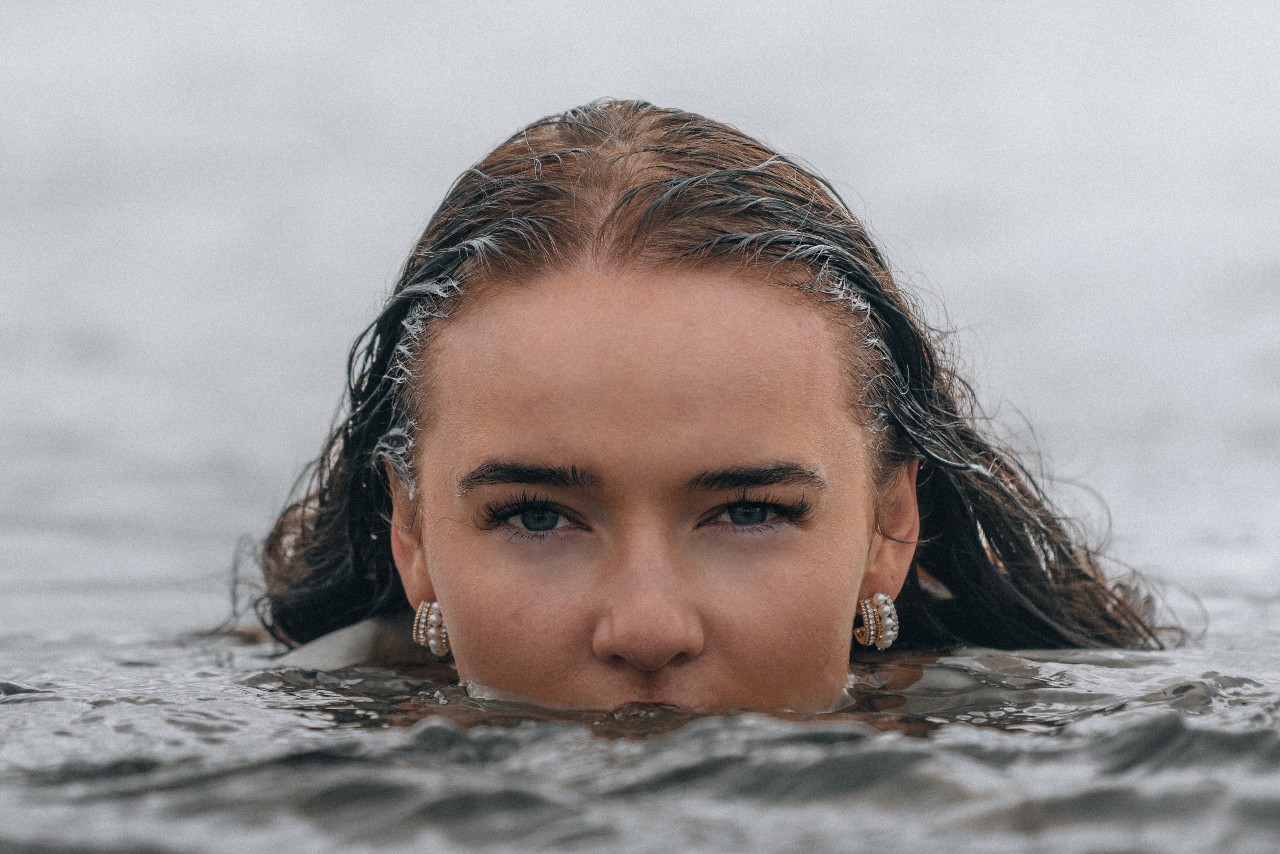 a woman whose face is halfway submerged in water, wearing pearl huggie earrings