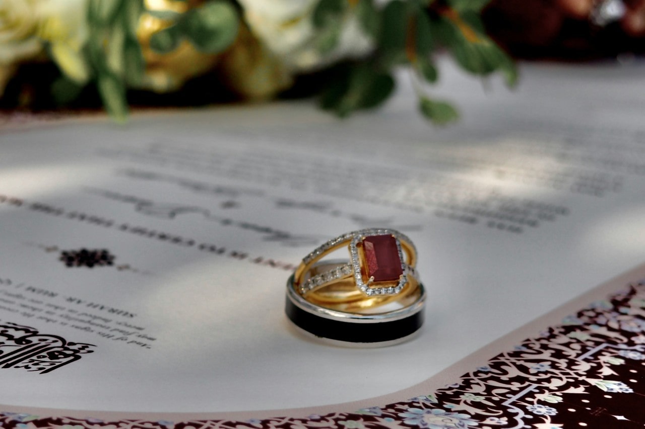 A close-up of wedding rings, including a gold and silver engagement ring with a red gemstone, resting on a marriage certificate with a blurred floral background