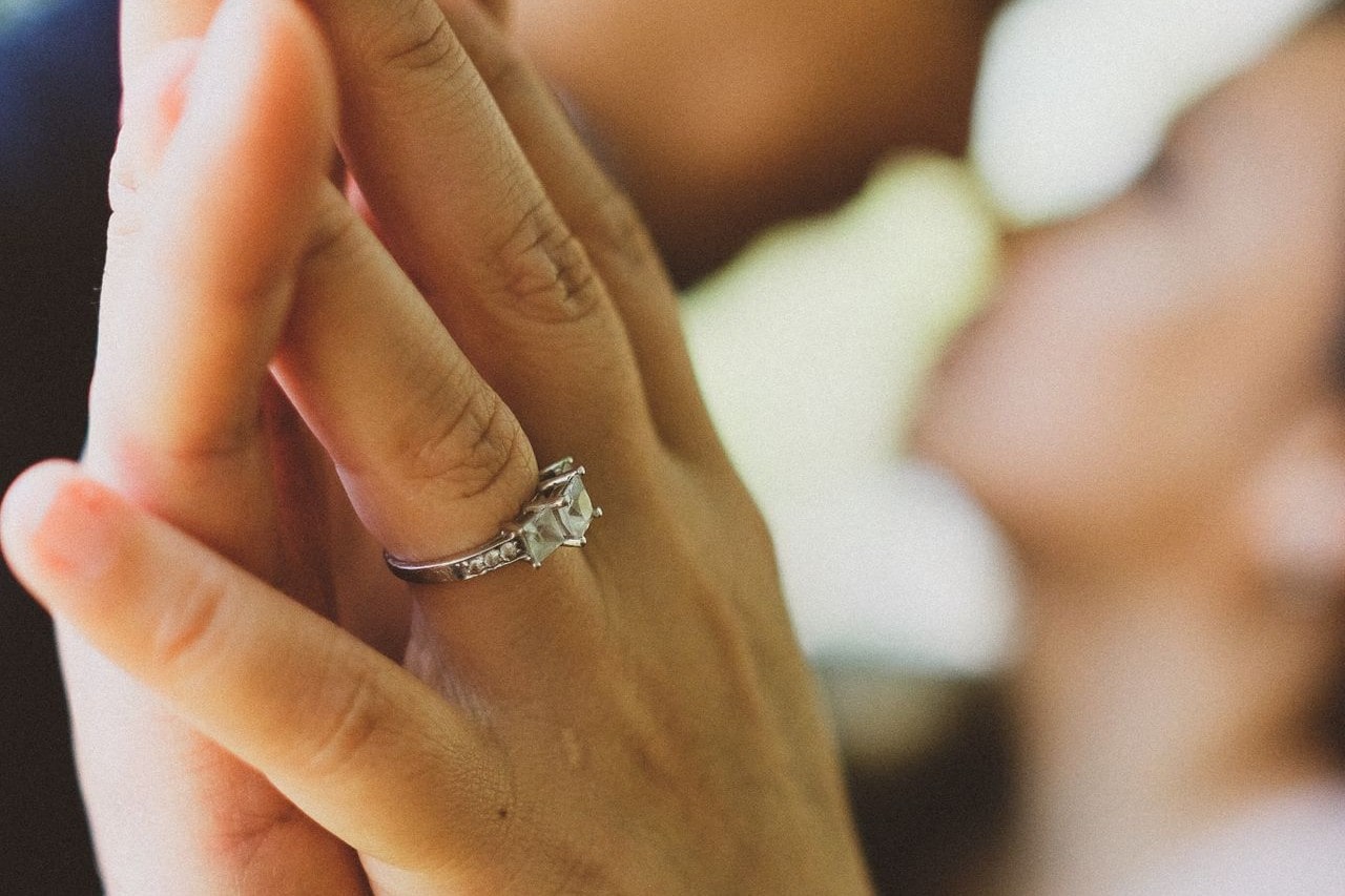 A couple holding hands, highlighting a silver engagement ring with a square-cut gemstone against a softly blurred background