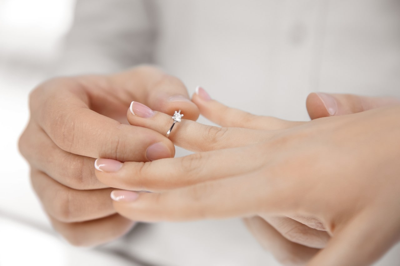 A close-up of groom placing a classic solitaire engagement ring, featuring a single round-cut diamond on a sleek silver band, onto bride’s finger