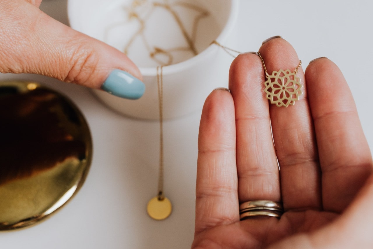 A close-up of a woman’s hands as she selects a gold necklace from her collection.